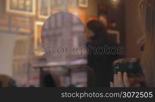 Young woman with digital camera taking shots of a boyfriend or friend in cafe, then they looking at photos on display. View through the window