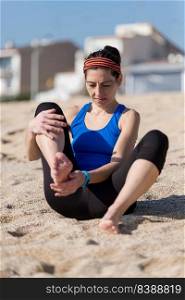 Young woman with headband and activewear sitting on beach sand, cleaning barefoot
