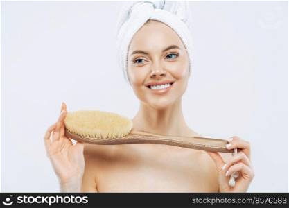 Young woman with healthy fresh skin, uses body brush, smiles gently, wears bath towel on head, poses topless, isolated over white background. Spa accessories. Women, body care, hygiene concept