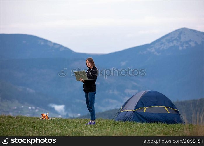 Young woman with laptop. Background beautiful mountains . Tourist girl is standing on a mountain near a tent, having dinner, burning fire.. Young woman with laptop. Background beautiful mountains . Tourist girl is standing on a mountain near a tent, having dinner, burning fire