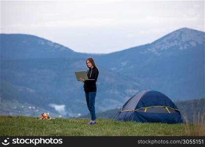 Young woman with laptop. Background beautiful mountains . Tourist girl is standing on a mountain near a tent, having dinner, burning fire.. Young woman with laptop. Background beautiful mountains . Tourist girl is standing on a mountain near a tent, having dinner, burning fire