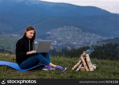 Young woman with laptop in the mountains. Girl works while sitting on the grass, the bonfire is lit on the side Work, business, freelance. Place for inscription. Young woman with laptop in the mountains. Girl works while sitting on the grass, the bonfire is lit on the side Work, business, freelance. Place for inscription.