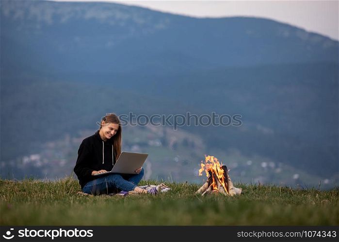 Young woman with laptop in the mountains. Girl works while sitting on the grass, the bonfire is lit on the side. Work, business, freelance. Place for inscription. Young woman with laptop in the mountains. Girl works while sitting on the grass, the bonfire is lit on the side. Work, business, freelance. Place for inscription.