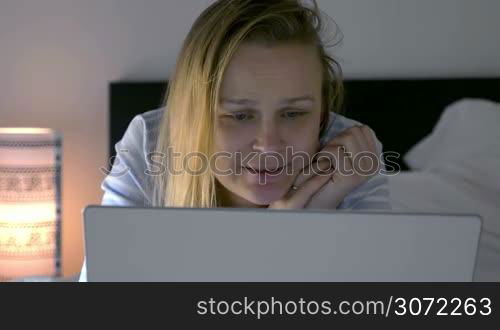 Young woman with laptop lying in bed in the evening. She watching movie triggering emotions of sadness and laugh