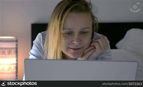 Young woman with laptop lying in bed in the evening. She watching movie triggering emotions of sadness and laugh