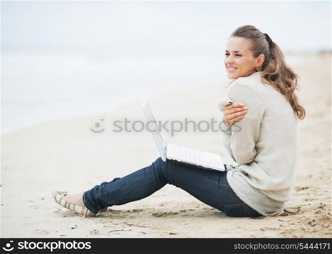 Young woman with laptop wrapping in sweater while sitting on lonely beach and