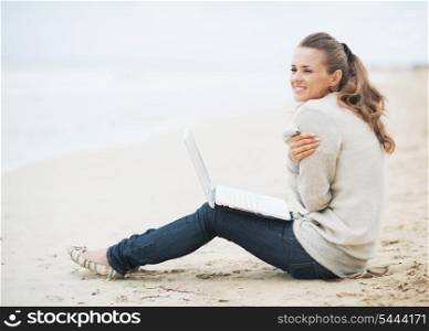Young woman with laptop wrapping in sweater while sitting on lonely beach and