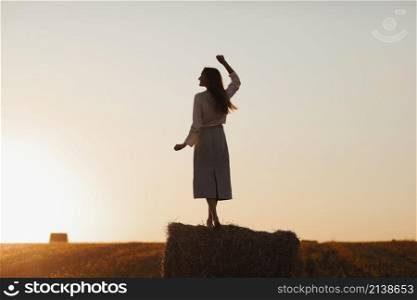 Young woman with long hair is standing and having fun on straw bale in field in summer on sunset. Female portrait in natural rural scene. Environmental eco tourism concept. Young woman with long hair is standing and having fun on straw bale in field in summer on sunset. Female portrait in natural rural scene. Environmental eco tourism concept.