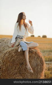 Young woman with long hair, wearing jeans skirt, light shirt and straw bag in hand, sitting on bale on field in summer. Female portrait in natural rural scene. Environmental eco tourism concept. Young woman with long hair, wearing jeans skirt, light shirt and straw bag in hand, sitting on bale on field in summer. Female portrait in natural rural scene. Environmental eco tourism concept.