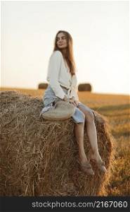 Young woman with long hair, wearing jeans skirt, light shirt and straw bag in hand, sitting on bale on field in summer. Female portrait in natural rural scene. Environmental eco tourism concept. Young woman with long hair, wearing jeans skirt, light shirt and straw bag in hand, sitting on bale on field in summer. Female portrait in natural rural scene. Environmental eco tourism concept.