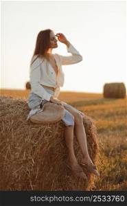 Young woman with long hair, wearing jeans skirt, light shirt and straw bag in hand, sitting on bale on field in summer. Female portrait in natural rural scene. Environmental eco tourism concept. Young woman with long hair, wearing jeans skirt, light shirt and straw bag in hand, sitting on bale on field in summer. Female portrait in natural rural scene. Environmental eco tourism concept.