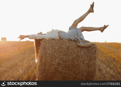 Young woman with long hair, wearing jeans skirt, light shirt is lying on straw bale in field in summer on sunset. Female portrait in natural rural scene. Environmental eco tourism concept. Young woman with long hair, wearing jeans skirt, light shirt is lying on straw bale in field in summer on sunset. Female portrait in natural rural scene. Environmental eco tourism concept.