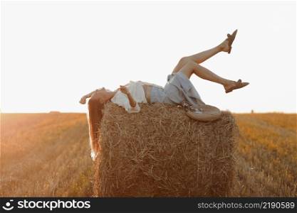 Young woman with long hair, wearing jeans skirt, light shirt is lying on straw bale in field in summer on sunset. Female portrait in natural rural scene. Environmental eco tourism concept. Young woman with long hair, wearing jeans skirt, light shirt is lying on straw bale in field in summer on sunset. Female portrait in natural rural scene. Environmental eco tourism concept.