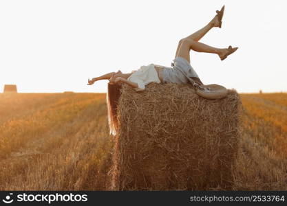 Young woman with long hair, wearing jeans skirt, light shirt is lying on straw bale in field in summer on sunset. Female portrait in natural rural scene. Environmental eco tourism concept. Young woman with long hair, wearing jeans skirt, light shirt is lying on straw bale in field in summer on sunset. Female portrait in natural rural scene. Environmental eco tourism concept.