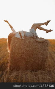 Young woman with long hair, wearing jeans skirt, light shirt is lying on straw bale in field in summer on sunset. Female portrait in natural rural scene. Environmental eco tourism concept. Young woman with long hair, wearing jeans skirt, light shirt is lying on straw bale in field in summer on sunset. Female portrait in natural rural scene. Environmental eco tourism concept.