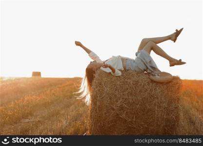 Young woman with long hair, wearing jeans skirt, light shirt is lying on straw bale in field in summer on sunset. Female portrait in natural rural scene. Environmental eco tourism concept. Young woman with long hair, wearing jeans skirt, light shirt is lying on straw bale in field in summer on sunset. Female portrait in natural rural scene. Environmental eco tourism concept.