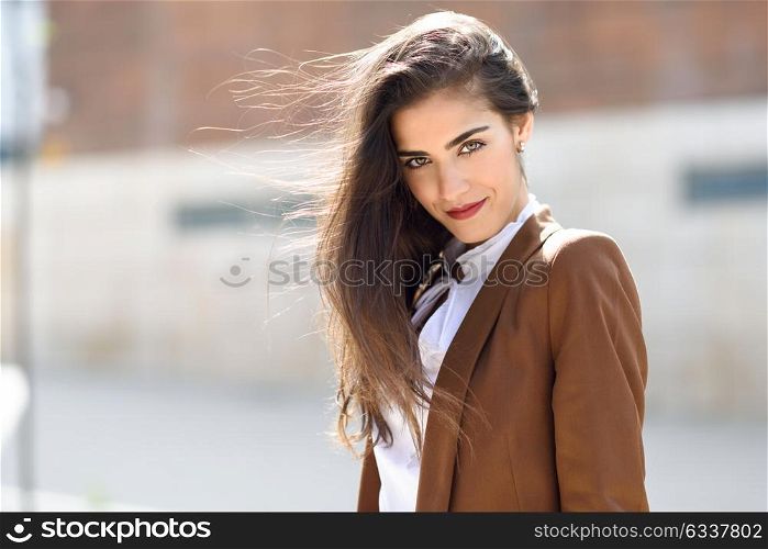 Young woman with nice hair in the wind standing in urban background. Businesswoman wearing formal wear with wavy hairstyle. Young girl with brown jacket and trousers.