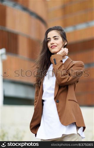 Young woman with nice hair standing outside of office building. Businesswoman wearing formal wear with wavy hairstyle. Young girl with brown jacket and trousers.