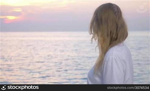 Young woman with pink ribbon by the sea at sunset. She turning to camera showing breast cancer awareness sign on her t-shirt. National Breast Cancer Awareness Month