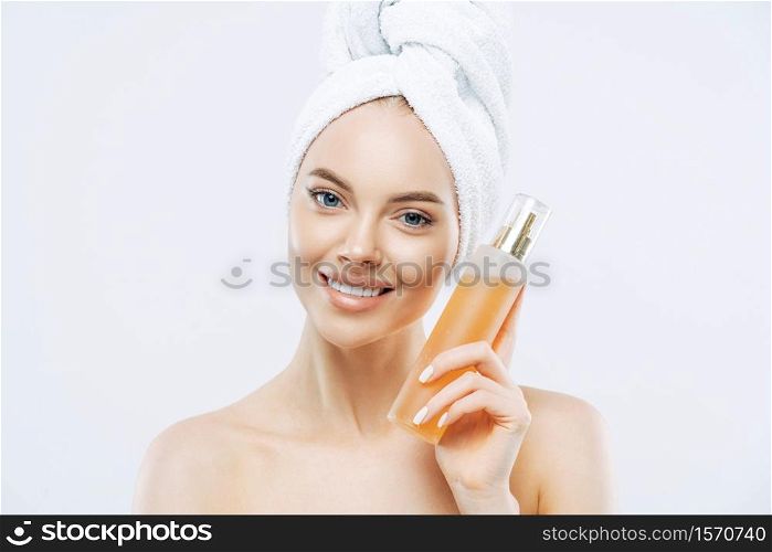 Young woman with positive smile, holds big bottle of aromatic parfum with flower odour, has well cared complexion, healthy skin, wears bath towel on head, stands shirtless isolated on white background