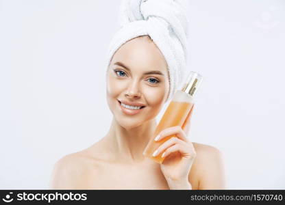 Young woman with positive smile, holds big bottle of aromatic parfum with flower odour, has well cared complexion, healthy skin, wears bath towel on head, stands shirtless isolated on white background