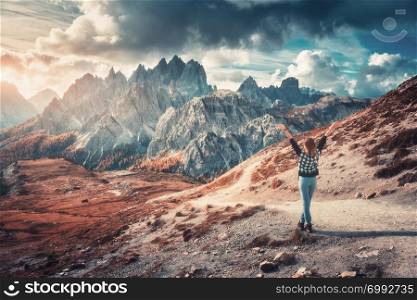 Young woman with raised up arms and high mountains at sunset in autumn in Dolomites, Italy. Landscape with happy girl, dramatic sky with clouds, orange trees, high rocks in italian alps in fall