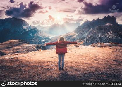 Young woman with raised up arms and mountains at sunset in autumn in Dolomites, Italy. Landscape with happy girl in red jacket, dramatic sky with clouds, orange grass and trees, high rocks in fall