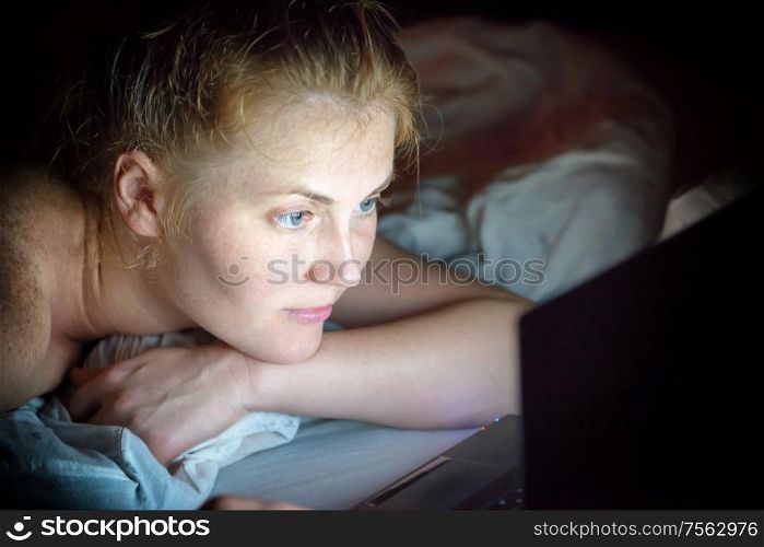 Young woman with red hair lying in bed at night with laptop and searching internet