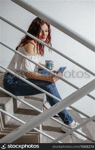 Young woman with red hair sitting on stairs and speaking on her smartphone and drinking coffee.