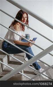 Young woman with red hair sitting on stairs and speaking on her smartphone and drinking coffee.