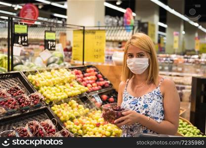 Young woman with shopping cart wears medical mask and buys cherries fruits. Shopping during the Coronavirus Covid-19 epidemic 2020. Young girl buys fruits at store or supermarket