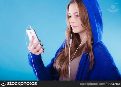 young woman with smart phone listening music. Teen stylish long hair girl in hood relaxing or learning language Studio shot on blue.