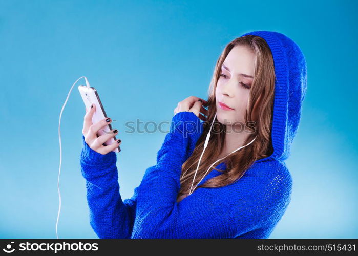 young woman with smart phone listening music. Teen stylish long hair girl in hood relaxing or learning language. Studio shot on blue.