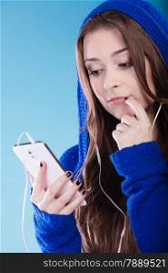 young woman with smart phone listening music. Teen stylish long hair girl in hood relaxing or learning language Studio shot on blue.