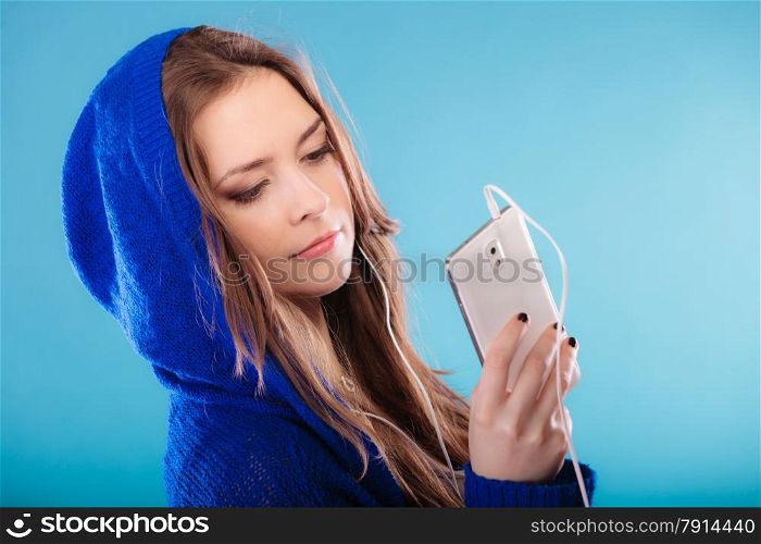 young woman with smart phone listening music. Teen stylish long hair girl in hood relaxing or learning language Studio shot on blue.
