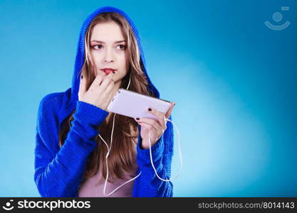young woman with smart phone listening music. Teen stylish long hair girl in hood relaxing or learning language. Studio shot on blue.