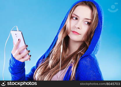 young woman with smart phone listening music. Teen stylish long hair girl in hood relaxing or learning language. Studio shot on blue.