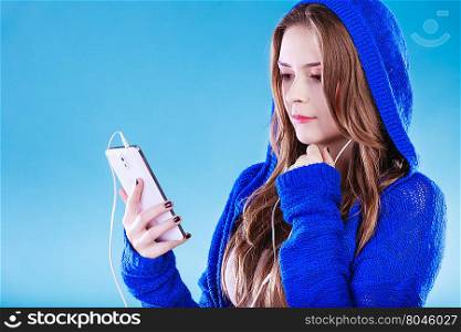 young woman with smart phone listening music. Teen stylish long hair girl in hood relaxing or learning language. Studio shot on blue.