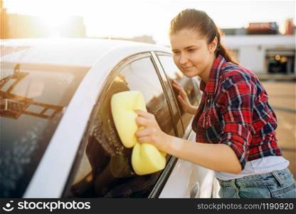 Young woman with sponge scrubbing vehicle glass with foam, car wash. Lady on self-service automobile washing. Outdoor carwash. Young woman scrubbing vehicle glass with foam