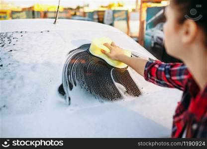 Young woman with sponge scrubbing vehicle glass with foam, car wash. Lady on self-service automobile washing. Outdoor carwash. Young woman scrubbing vehicle glass with foam