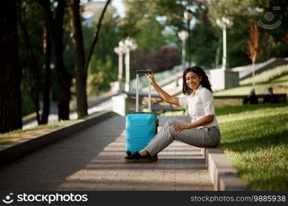 Young woman with suitcase sitting on walkway in summer park. Female traveler with luggage leisures outdoors, passenger with bag resting in nature. Girl with baggage relax on city alley. Woman with suitcase sitting on walkway in park