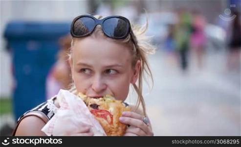 Young woman with sunglasses on her head eating fast food in the street and looking around being thoughtful.