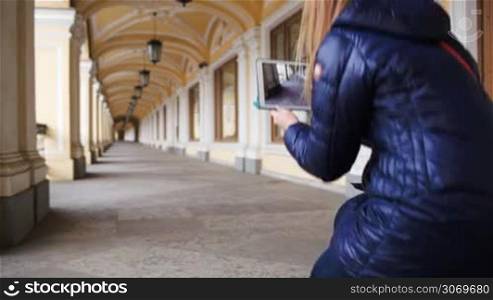 Young woman with touchpad hunkering down to make a shot of classic style hall in perspective, then she going away