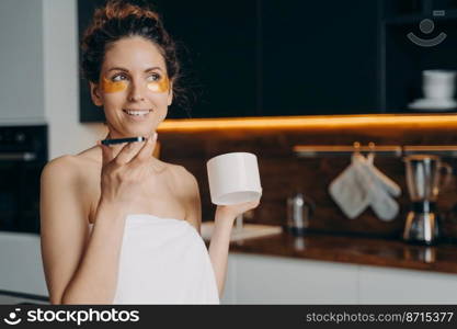 Young woman with under eye patches sends audio voice message holding smartphone and coffee cup on kitchen. Hispanic girl wrapped in towel talking on phone during skincare procedure after shower.. Hispanic girl sends voice message holding smartphone, coffee cup during skincare treatment at home