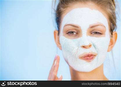 Young woman with white dry mud mask on face, against blue. Teen girl checking result of treatment. Skincare.. Girl with dry white mud mask on face