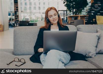 Young woman working as call center agent online, communicates with customers using wireless earphones headset and laptop, working remotely on support hotline while sitting on couch in coffee house. Young woman working as call center agent online using wireless earphones headset and laptop