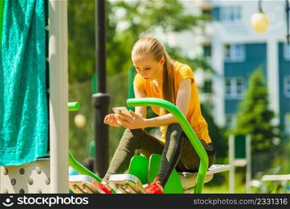 Young woman working out in outdoor gym. Girl holding mobile phone while doing exercises on street machine.. Girl doing exercises outdoor, holding phone
