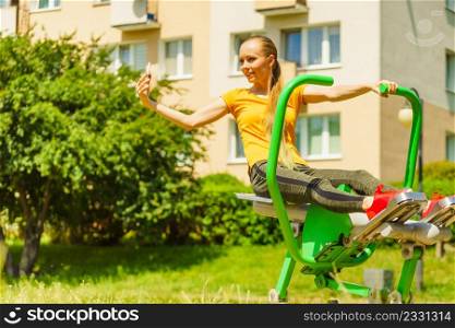 Young woman working out in outdoor gym. Girl taking selfie photo with smartphone while doing exercises on street machine.. Girl doing exercises outdoor, taking selfie