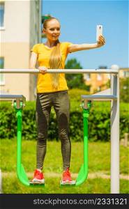 Young woman working out in outdoor gym. Girl taking selfie photo with smartphone while doing exercises on street machine.. Girl doing exercises outdoor, taking selfie