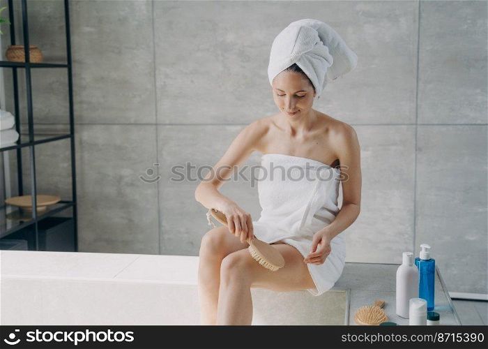 Young woman wrapped in towel massages hip with natural massaging brush, sitting on the side of bathtub in modern bathroom after the shower. Cellulite treatment, skincare, body care concept.. Woman wrapped in towel massages hip with massaging brush in bathroom. Cellulite treatment, body care